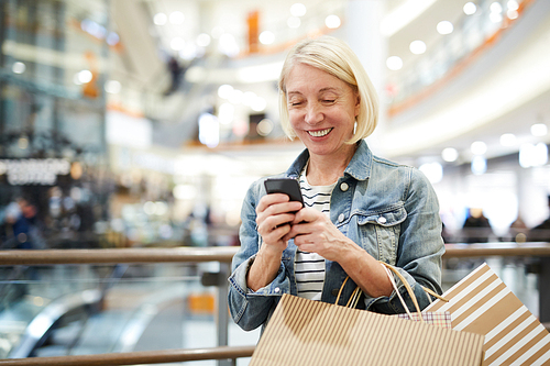 Happy excited mature woman with short blond hair standing in corridor of shopping mall and texting sms on smartphone while chatting online with shopping assistant
