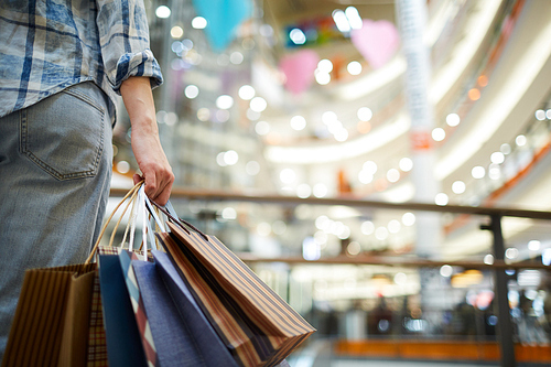 Close-up of unrecognizable woman in casual clothing standing in spacious shopping mall with lights and holding many paper bags