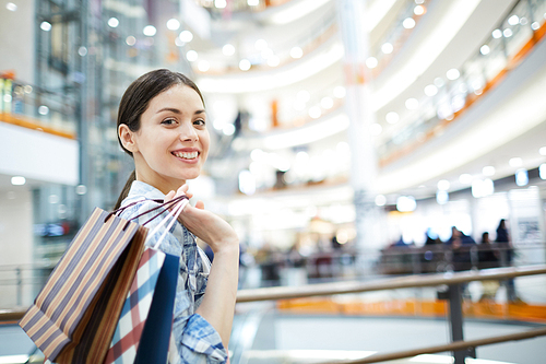 Positive pretty girl with black hair standing in modern shopping mall and holding paper bags with purchases on shoulder, she 