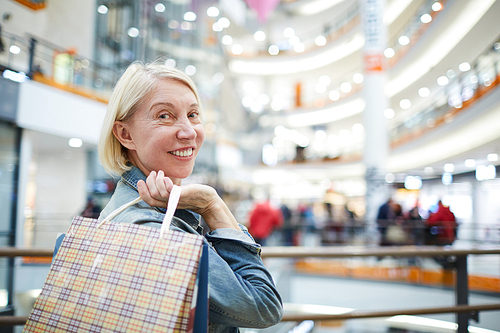 Positive confident attractive mature lady with short blond hair standing in entrance hall of shopping mall and holding bags on shoulder, she enjoying good shopping