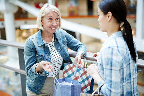 Smiling satisfied mature woman frowning forehead from excitement showing purchase in shopping bag to friend while they chatting in lobby of mall