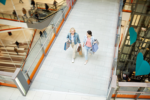 Directly above view of happy excited mother and daughter in casual outfits carrying many shopping bags and talking while walking over mall