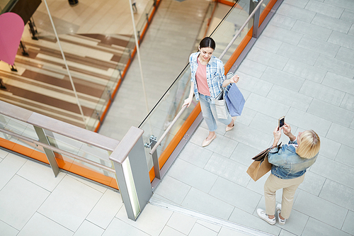 Top view of blond lady using smartphone to photograph friend with shopping bags in modern mall