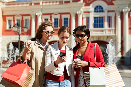 Content curious young multi-ethnic female friends standing outside on street with fountains and using shopping app on smartphone together