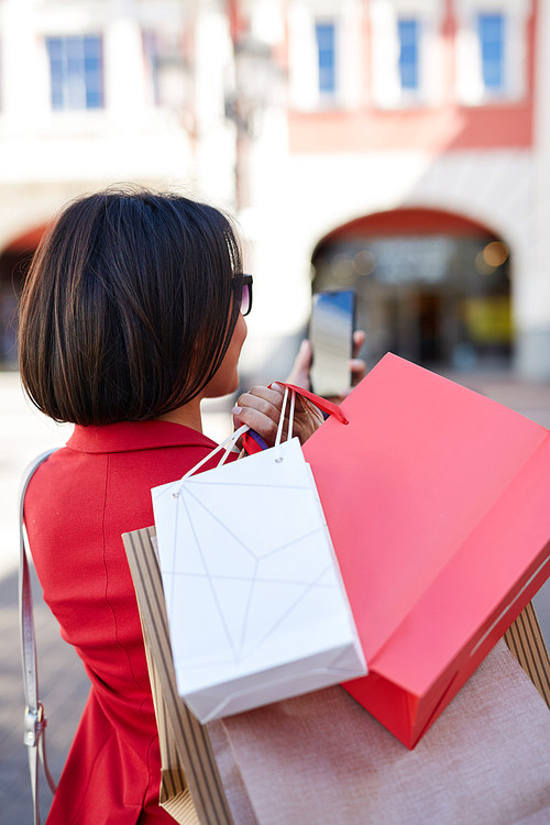 Rear view of brunette black woman in red jacket taking fashion selfie with heap of shopping bags outdoors