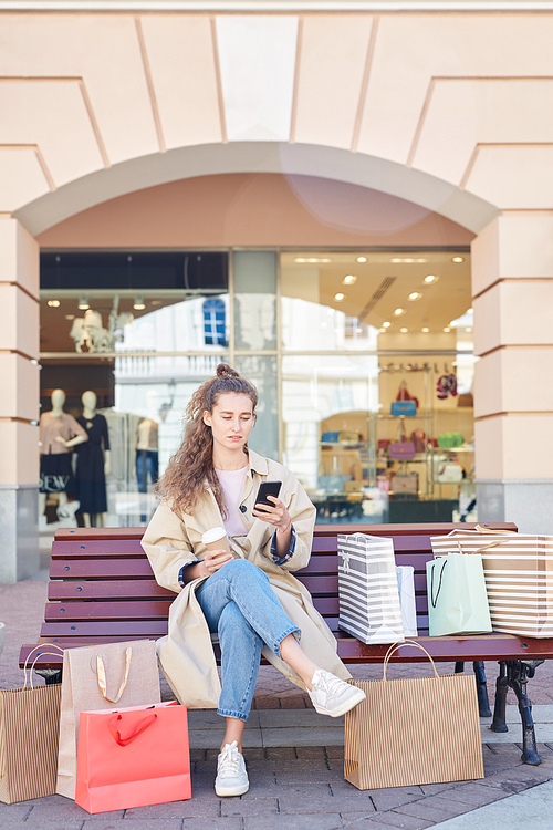 Serious stylish girl with long curly hair sitting on wooden bench with shopping bags and using smartphone while browsing website with trendy outfits