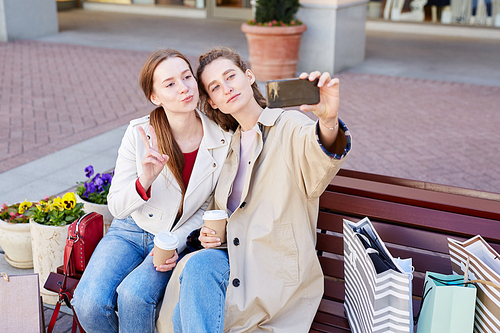 Content stylish young women in casual outfits sitting on bench with coffee cups and posing for selfie made on smartphone