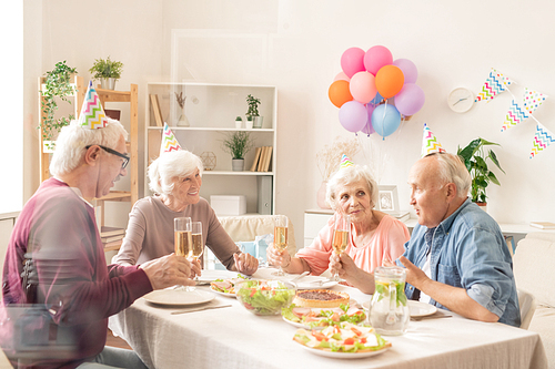 Four senior friends with champagne sitting by festive table during home birthday party