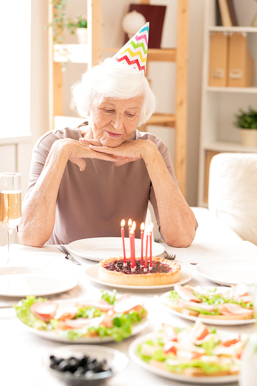 Aged retired female looking at burning candles on homemade cake while sitting by served birthday table
