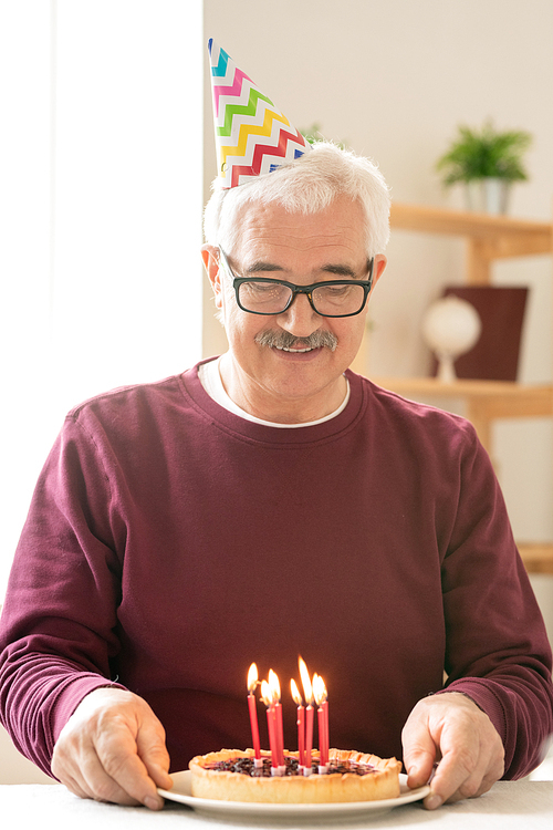 Happy senior man making birthday wish while looking at burning candles on homemade cake