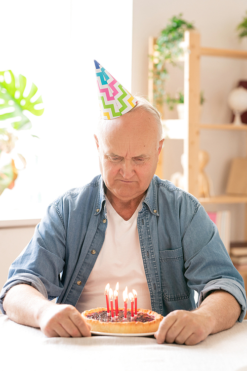 Retired man in birthday cap making wish in front of cake with candles by table