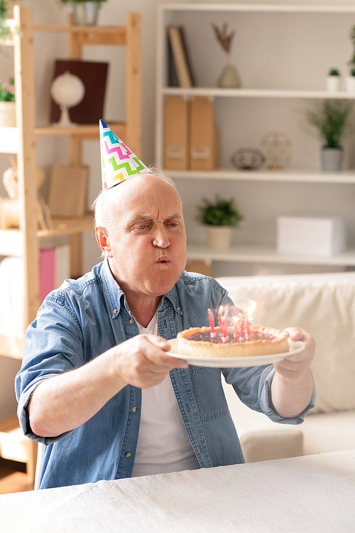 Casual senior man holding plate with birthday cake in front of him and blowing candles