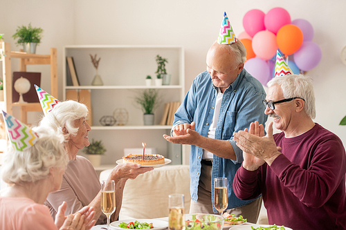 Senior friends clapping hands while looking at their friend with birthday cake by served table