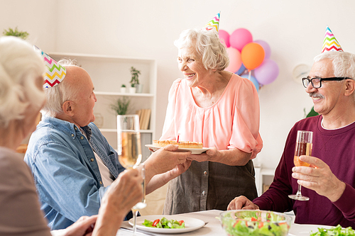 Cheerful aged female with birthday cake looking at one of friends toasting with champagne at home party