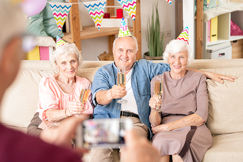 Group of aged friendly people toasting for birthday while sitting on couch at home part