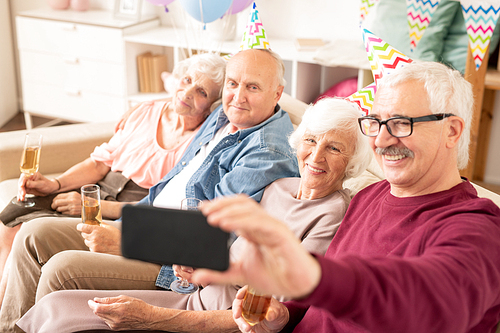 Group of cheerful seniors making selfie on couch while gathered for birthday party at home