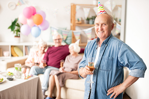 Smiling senior man with flute of champagne in front of camera on background of his friends