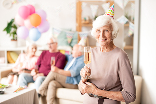 Cheerful grandma in birthday cap holding flute of champagne while standing in front of camera