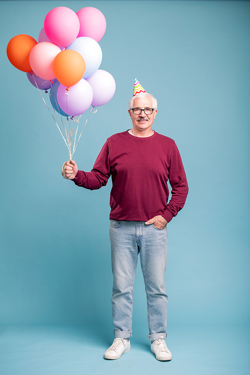 Happy mature man in birthday cap and casualwear holding balloons while posing on blue background