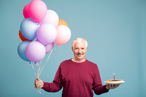 Happy senior man with bunch of balloons and homemade birthday cake posing in studio