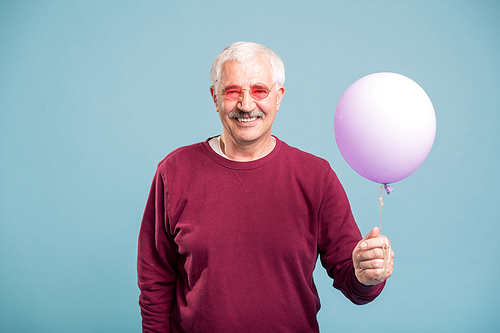Happy retired man in pullover and eyeglasses holding balloon while posing in studio