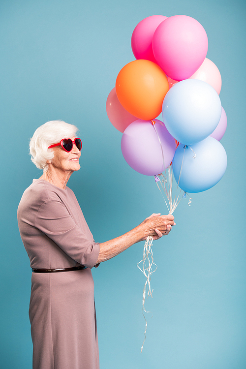 Elegant and gracious senior female holding bunch of colorful balloons while standing in studio