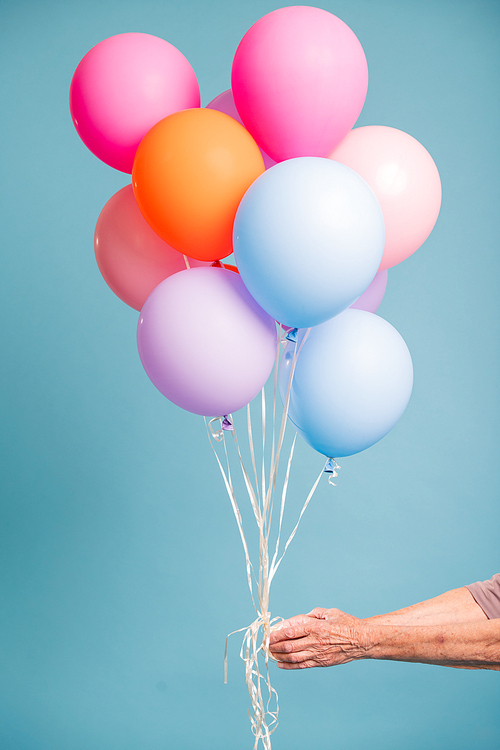 Hand of senior woman holding colorful balloons over blue background in isolation
