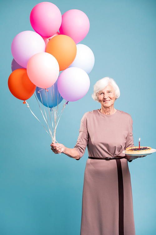 Happy aged woman in elegant dress holding pie with candle and bunch of colorful balloons