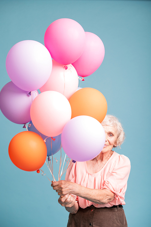 Happy senior woman peeking out of bunch of colorful balloons in isolation