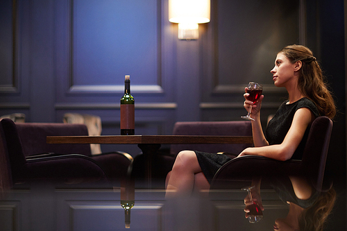 Pretty young woman in black dress sitting in armchair by table and having glass of red wine alone