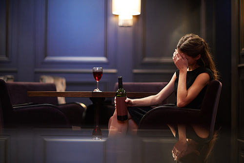 Lonely young woman with bottle of red wine sitting in armchair by table in luxurious restaurant with palm on face
