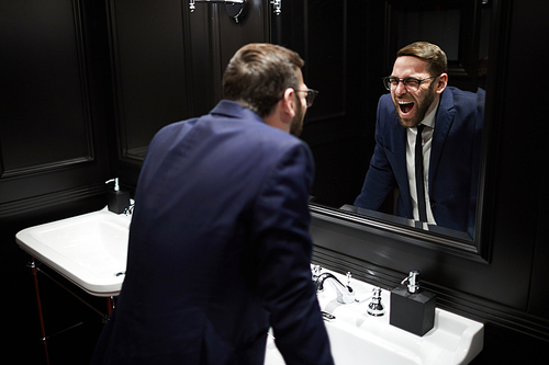 Young well-dressed man bending over sink in front of mirror and screaming while looking at himself