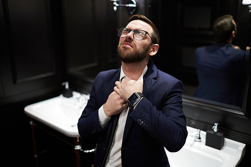 Contemporary businessman in suit keeping his hands on throat while loosing his necktie in lavatory