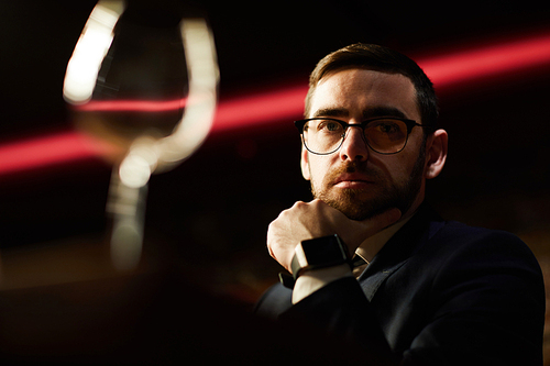 Serious businessman in eyeglasses keeping his hand by chin while sitting in luxurious restaurant