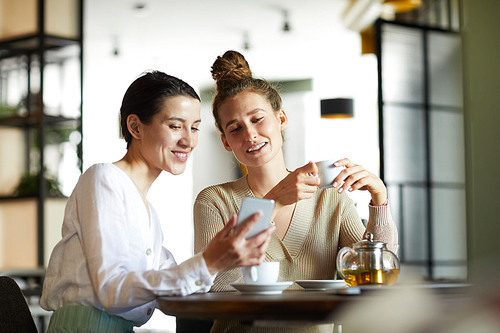 Two young females in casualwear watching curious video in smartphone while having tea in cozy cafe