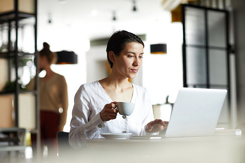Young serious businesswoman in white shirt having tea in front of laptop while organizing work or analyzing online data