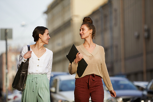 Two young happy urban women in casualwear chatting on their way to work or while taking walk along city road