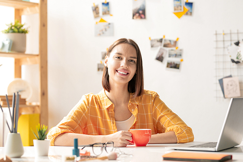 Happy brunette girl in casualwear looking at you with toothy smile while sitting by desk in office, having drink and networking