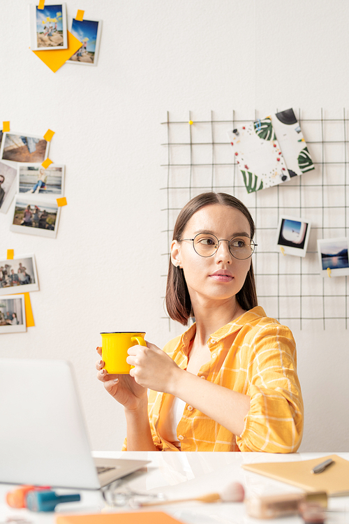 Young serious pretty businesswoman or designer in yellow casual shirt having tea or coffee while sitting by desk in front of laptop