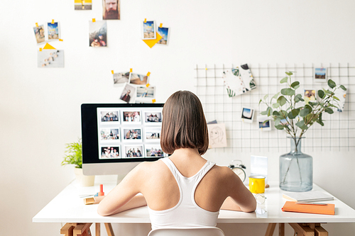 Back view of young female designer or editor in casualwear sitting in front of monitor while working over new collection of photos