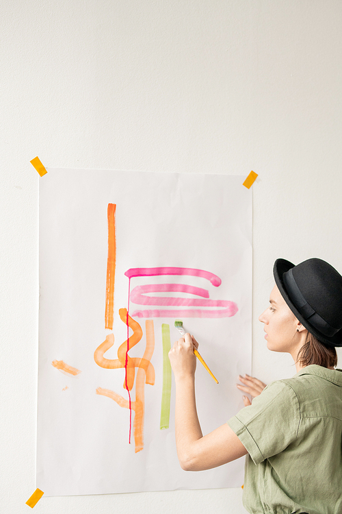 Young elegant female artist with paintbrush standing by sheet of paper on wall while painting with pink, yellow and orange watercolors