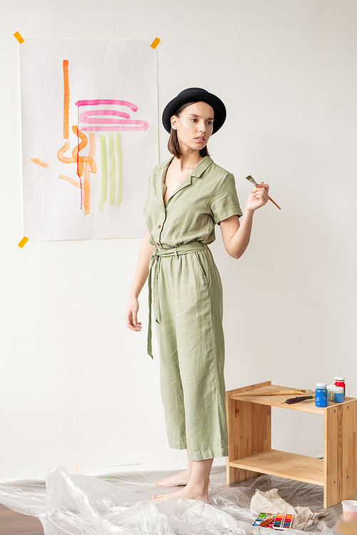 Young creative female in casualwear and black hat holding paintbrush while standing by wall and painting something abstract