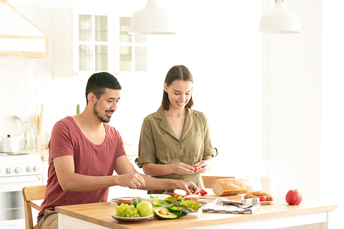 Happy young vegetarians in casualwear standing by kitchen table while preparing fresh vegetable salad or sandwiches