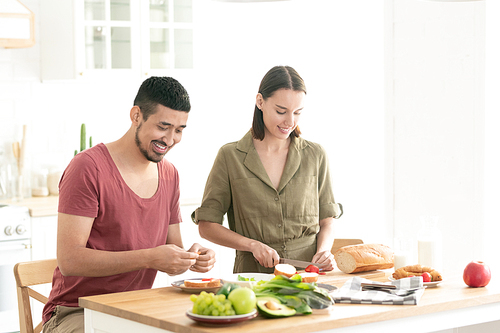 Happy young woman cutting fresh tomatoes on chopping board while her husband making vegetable sandwiches