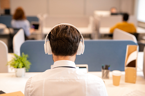 Rear view of office employee in wireless headphones sitting in modern office and working with customers online