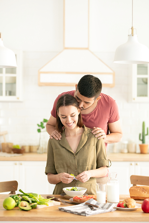 Young amorous husband giving his wife kiss on her head while she cooking vegetable salad for breakfast