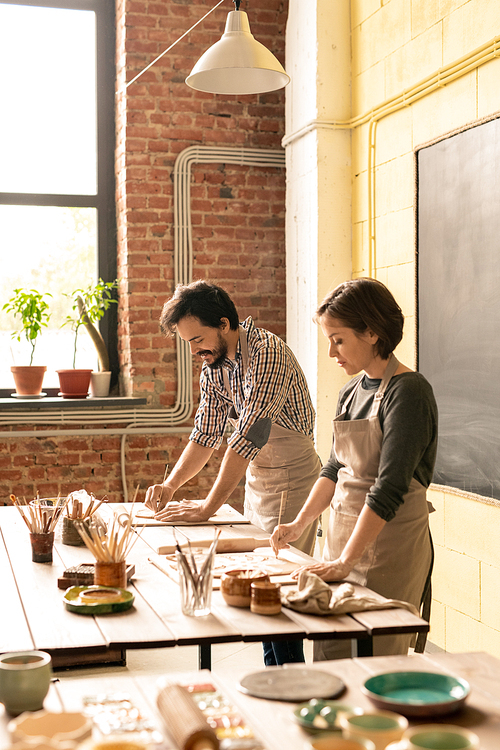 Young creative man and woman in workwear bending over wooden table while working over new collection of earthenware