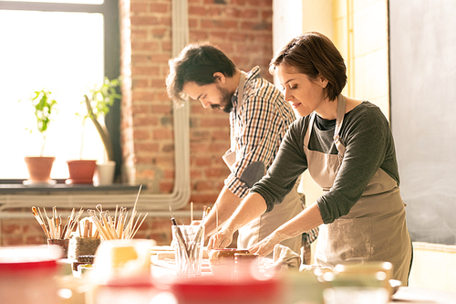 Young woman in apron and her colleague working over collection of clay items or earthenware in their own studio