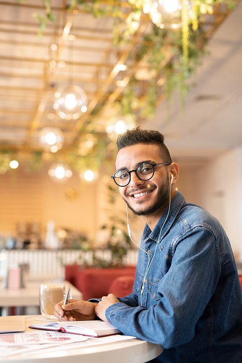 Happy guy in denim shirt organizing work while listening to music or audio lecture in earphones and making notes in notebook