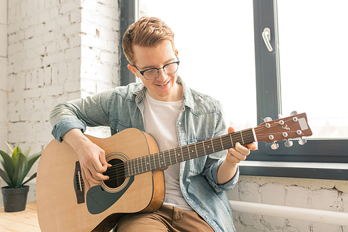 Happy guy in casualwear sitting by window and playing the guitar while singing at home party for his friends